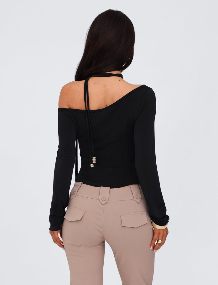 A woman with long, dark hair stands facing away from the camera. She is wearing the TIGERMIST MIETTA TOP in black, a long-sleeve, off-the-shoulder design featuring a multi-tie neck and high-stretch fabric. Paired with light beige, high-waisted pants with flap pockets, she exudes effortless elegance against a plain white background.