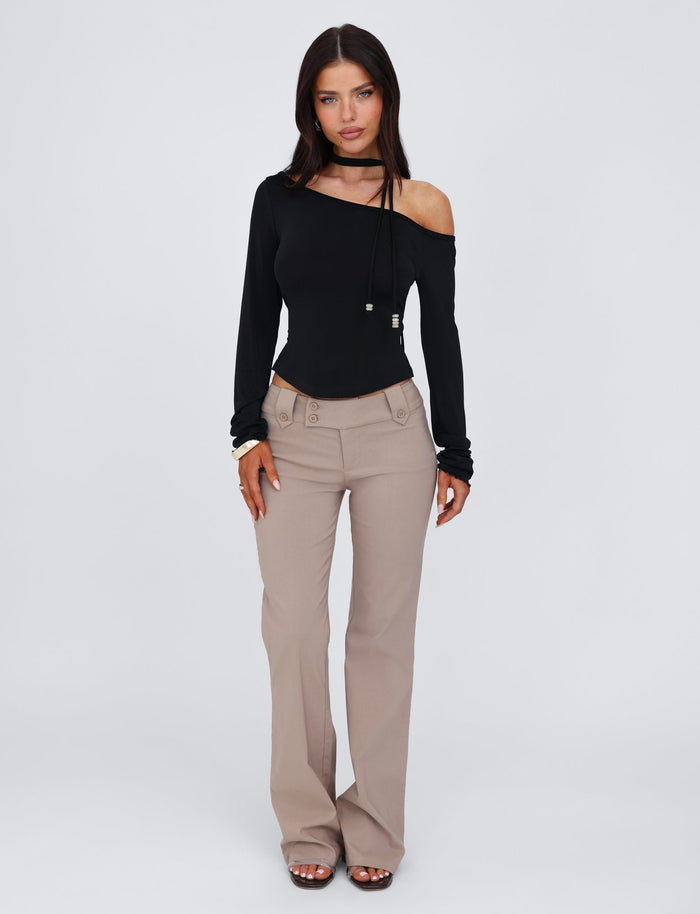 A woman is posed against a simple, light grey backdrop, showcasing a chic ensemble. She is dressed in the TIGERMIST MIETTA TOP - BLACK, which features an off-the-shoulder, long-sleeve design. Complementing her top are beige high-waisted, wide-leg pants. For footwear, she has opted for black open-toe heels and her long hair is styled straight.