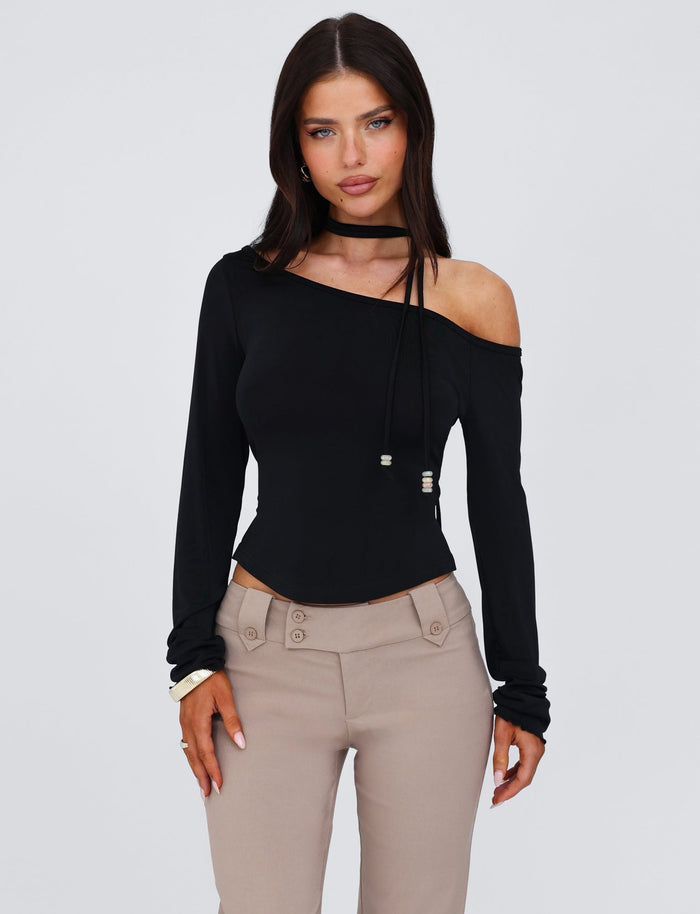 A woman with long dark hair is wearing the stylish TIGERMIST MIETTA TOP - BLACK, made from high-stretch fabric. The top features an off-the-shoulder design with long sleeves and is paired with taupe high-waisted pants. She has a neutral expression and her hands rest by her sides against a plain white background.