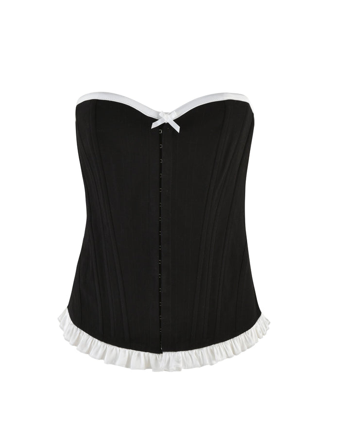 The TIGERMIST MILLIE CORSET 2.0 in black boasts vertical seams, white trim along the top edge, and a ruffled white trim along the bottom. This strapless corset features a small white bow at the top center and a front busk closure, offering a sleek bodycon fit.