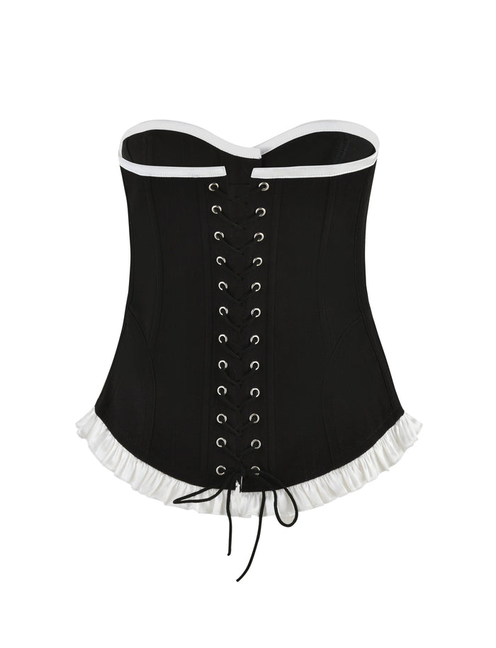 The TIGERMIST MILLIE CORSET 2.0 in black features a sweetheart neckline, front lacing with eyelets, and white trim. This bodycon fit corset is enhanced with a white ruffled hemline and black laces tied in a bow at the bottom.