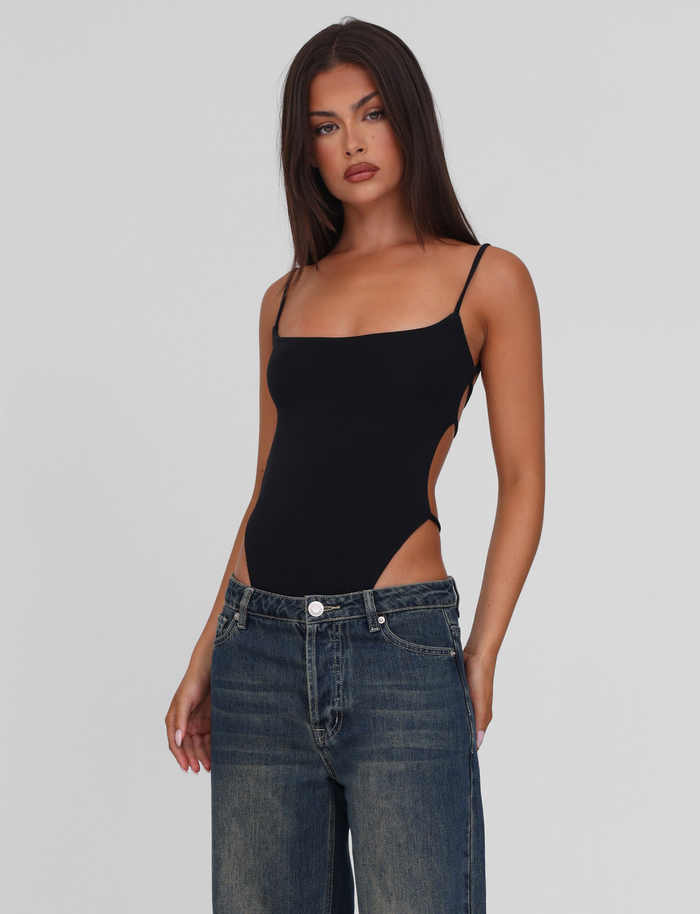 A woman with long dark hair models the TIGERMIST MINERVA BODYSUIT in black, featuring a backless design with thin straps and high-cut sides. She pairs it with loose blue jeans and poses against a plain light gray background, gazing slightly to the side.