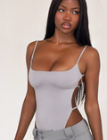 A woman with long straight hair wears the TIGERMIST MINERVA BODYSUIT in Frost Grey, featuring side cutouts and paired with matching pants. She gazes to the side with a neutral expression against a plain background.