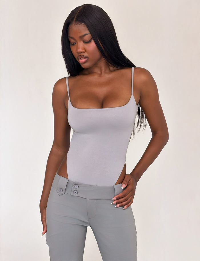 A woman with long straight black hair wears the TIGERMIST MINERVA BODYSUIT in Frost Grey and low-rise gray pants, standing against a plain light background while looking down to her right.