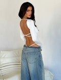 A woman with long, dark hair stands facing backward, turning her head slightly to the side. She is wearing the MINERVA LONG SLEEVE BODYSUIT - WHITE from TIGERMIST and high-waisted, loose-fitting blue jeans. She stands in front of a white wall and a beige textured couch.