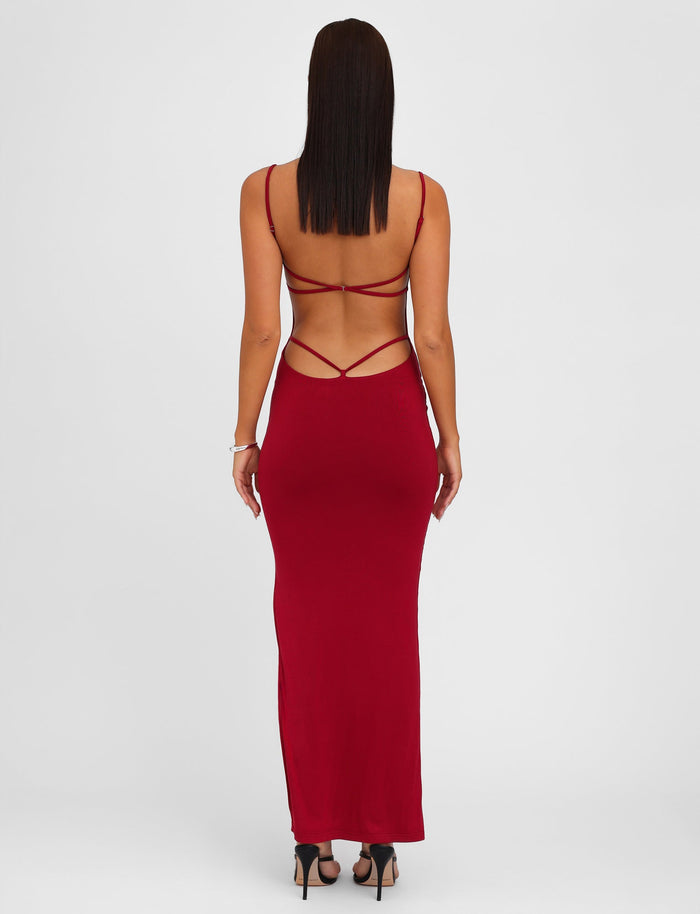 A woman faces away from the camera while wearing the TIGERMIST MINERVA MAXI DRESS in Berry Red—an elegant, long dress featuring a bodycon silhouette and adjustable shoulder straps. She has straight hair and complements her look with black high-heeled sandals against a plain white background.