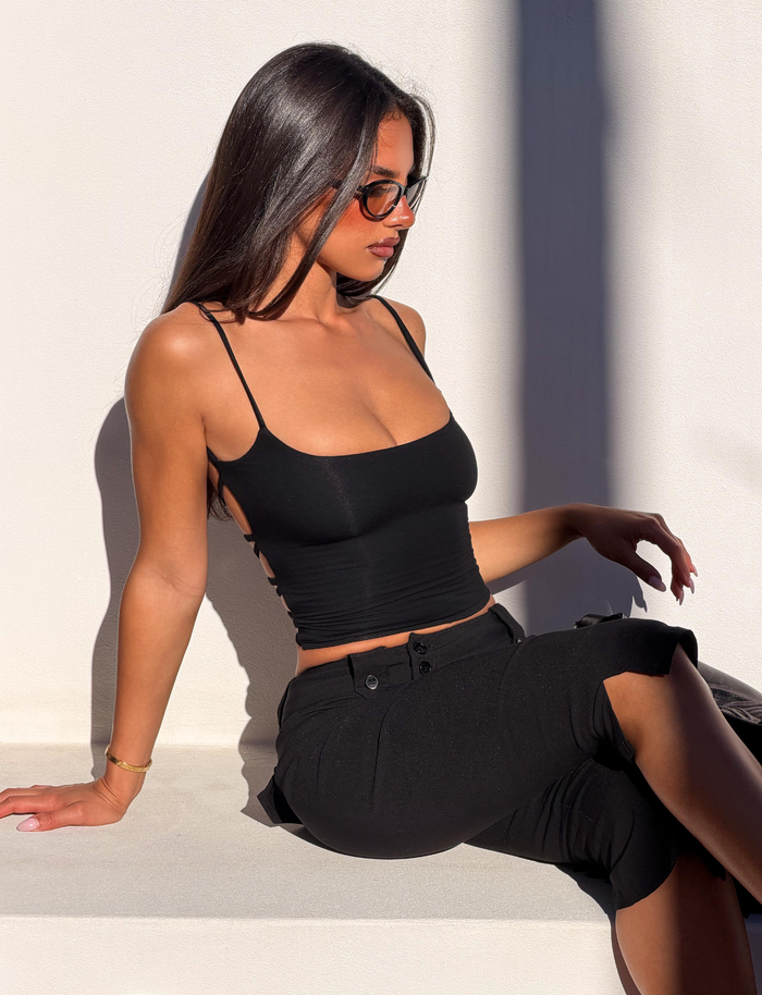 A woman with long dark hair and sunglasses wears the TIGERMIST MINERVA TOP - BLACK, styled with black pants, as she sits on a white surface in bright sunlight, casting bold shadows on the wall behind her.