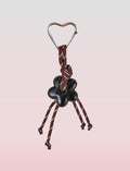 The Tigermist MIRIAM KEYCHAIN - CHOCOLATE features a heart-shaped metal carabiner clasp, attached to dark brown rope knotted decoratively. Four knotted ends dangle a black flower charm, set against a light pink background for soft contrast.