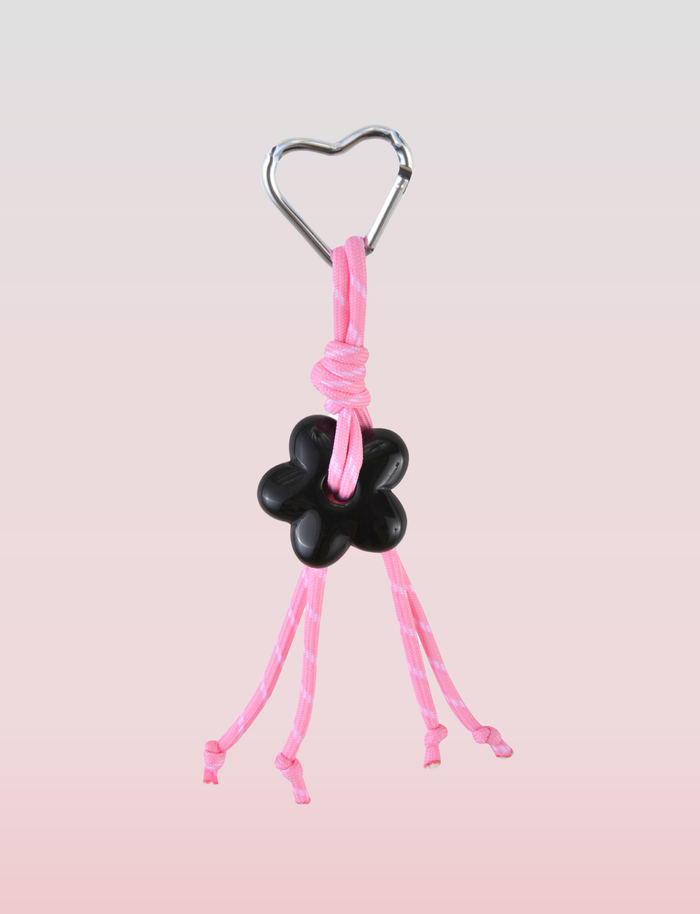 The TIGERMIST MIRIAM KEYCHAIN - HOT PINK features a heart-shaped metal ring, pink cord, black flower charm, and four loose pink cord ends on a light pink gradient background.