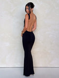 A woman poses with her back to the camera in a black TIGERMIST MORANA DRESS - BLACK, showcasing its stunning bodycon fit. Her elegant look, completed with black heels and an updo hairstyle, contrasts beautifully against the light textured background.