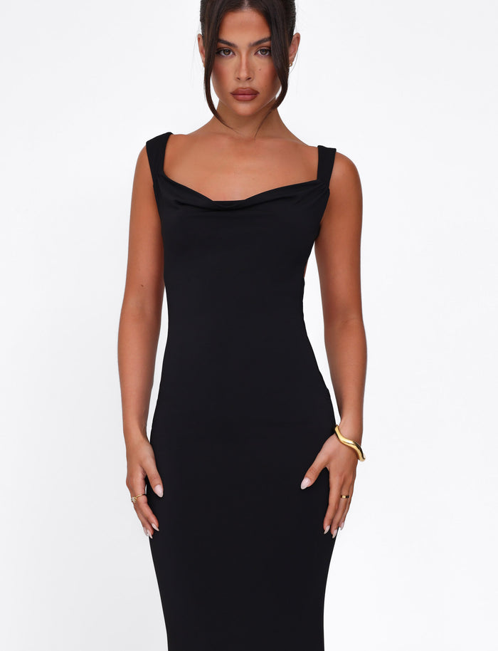 A person in the TIGERMIST MORANA DRESS - BLACK, a bodycon style, stands against a white backdrop with hands near hips adorned by a gold bracelet. Their hair is styled up, displaying a neutral expression.