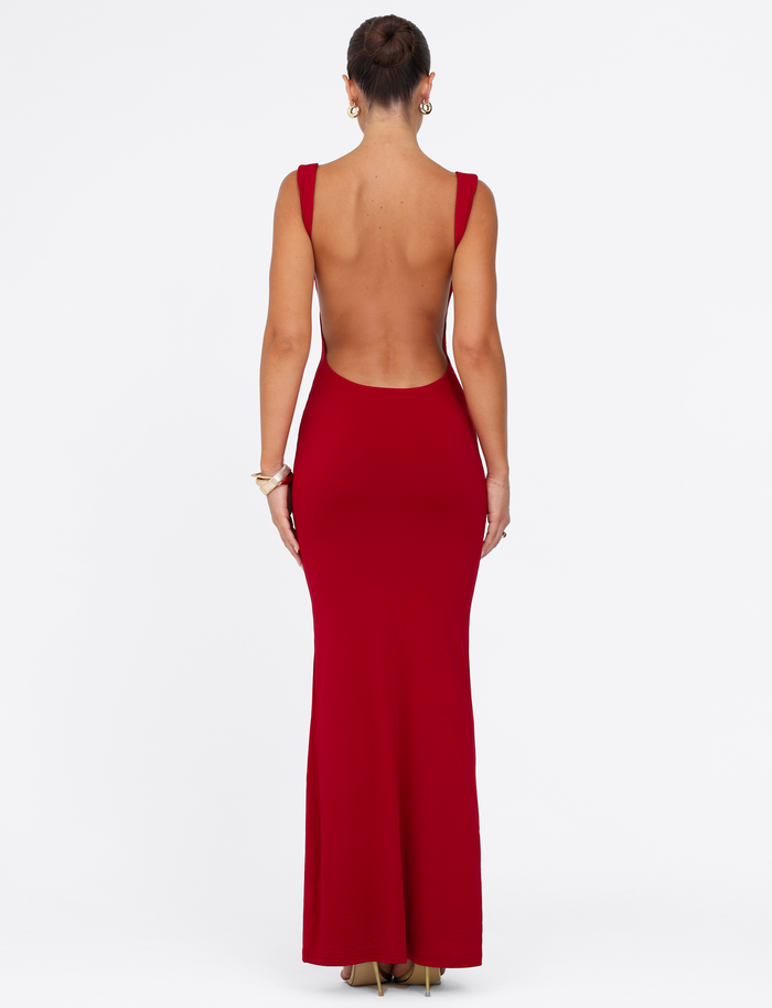 A woman in a sleeveless, backless TIGERMIST MORANA MAXI DRESS - RED, styled with gold heels, bracelets, and hoop earrings, her hair in a bun, poses against a plain white background.