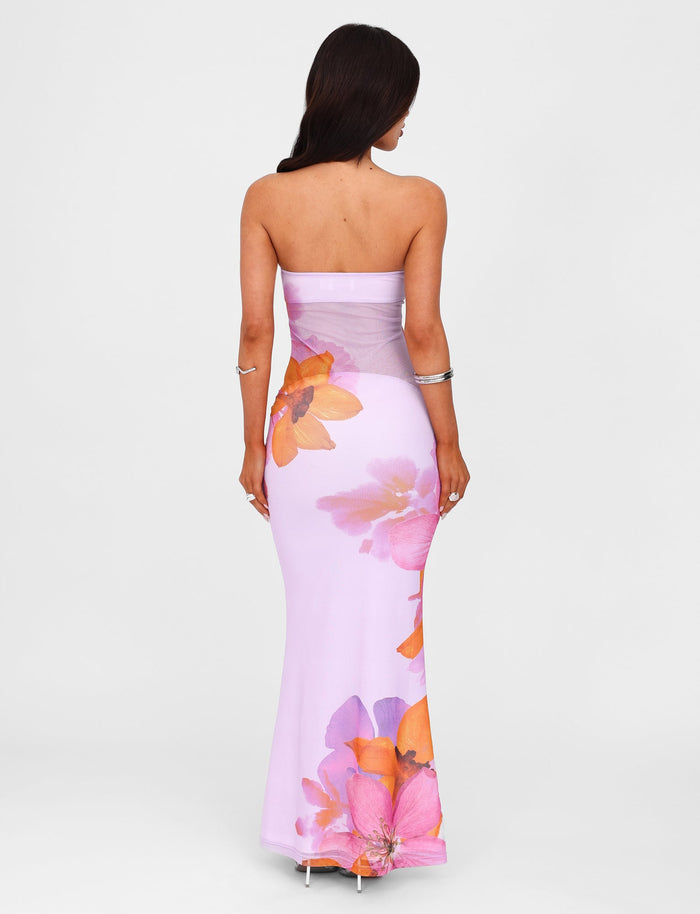 A woman with long dark hair stands with her back to the camera, wearing the MORGAN DRESS - PINK from the TIGERMIST x By Poppy Summer collection, featuring vibrant orange and pink flower prints. She has a silver bracelet on one wrist and is wearing high-heeled shoes against a plain white background.