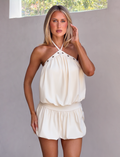 A woman with long blonde hair wears the sleeveless NATASHA DRESS in cream by TIGERMIST, featuring a halter neckline. She stands indoors against a light gray wall with sunlight streaming through the window above.