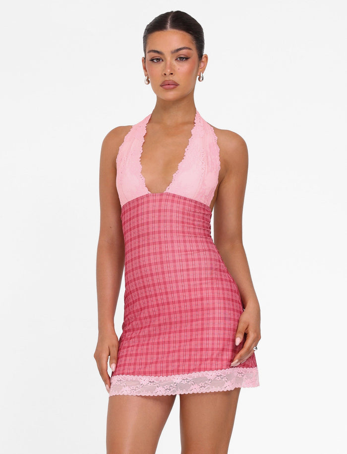 Against a white backdrop, a woman wears the NAVIA DRESS - RED CHECK from TIGERMIST, featuring a pink lace halter mini dress design with plaid patterns. Her hair is tied back, and she complements the look with elegant earrings.