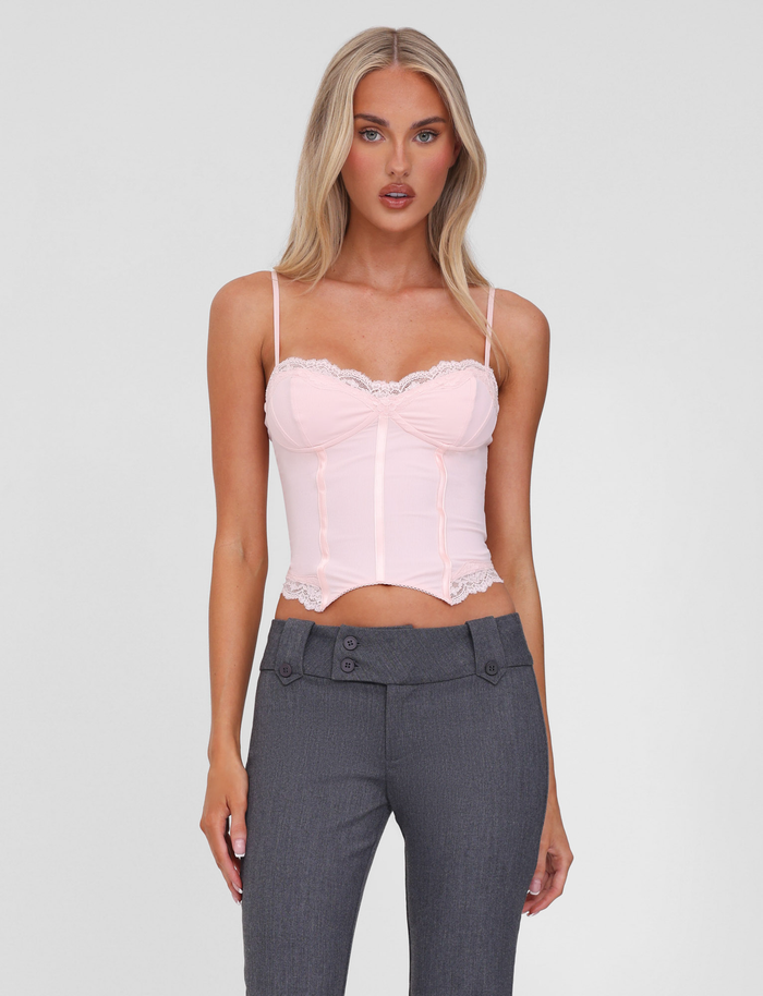 A woman with long blonde hair wears the TIGERMIST NILA CORSET TOP in soft pink with adjustable straps and high-waisted gray pants. She stands facing forward against a plain white background, her expression neutral.