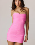 A woman with long brown hair wears the TIGERMIST NIYA BANDAGE MINI DRESS in pink, which features a strapless sweetheart neckline. She stands facing forward against a plain light background, displaying a small heart tattoo on her left shoulder.