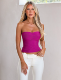 A woman with long blonde hair wears the TIGERMIST NIYA BANDAGE TOP in magenta and high-waisted white pants, standing by a light-colored wall with sunlight streaming through a window.
