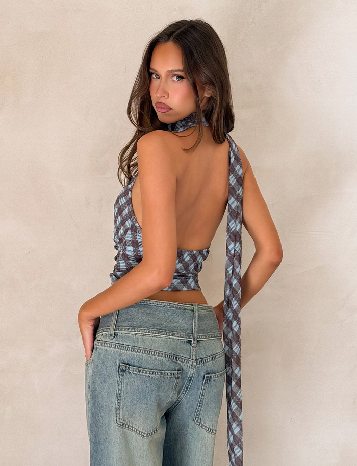A woman with long brown hair poses against a beige wall, wearing the TIGERMIST NOBLE TOP - BLUE CHECK with a plunge neckline and long tie, paired with light blue high-waisted jeans as she looks over her shoulder at the camera.