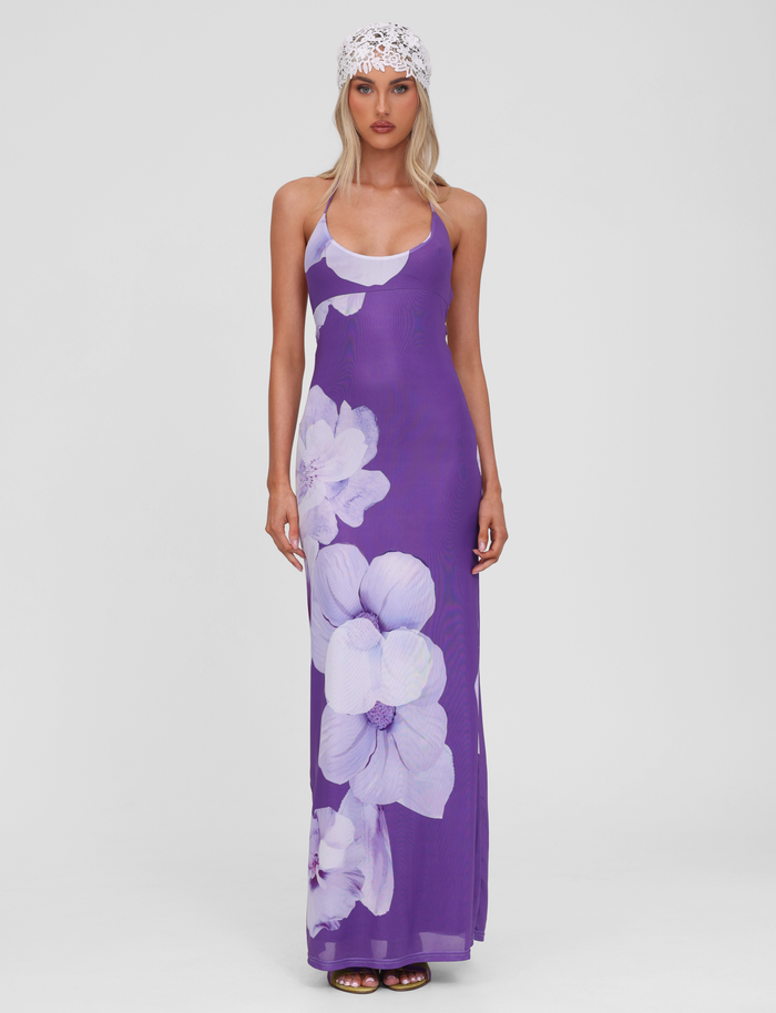 A woman with long blonde hair and a white lace headscarf faces forward, modeling the TIGERMIST NOUR MESH MAXI DRESS in Purple Macro Floral against a plain white background. The dress features a halter neckline and bold purple floral prints.