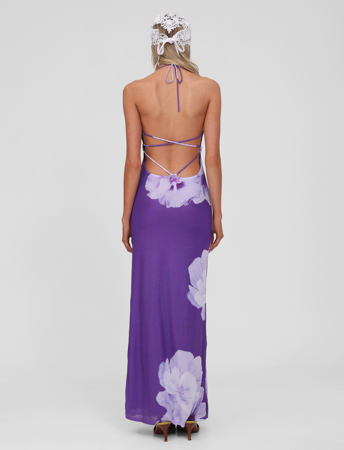 A woman with long blonde hair beneath a white crocheted head covering faces away, wearing the TIGERMIST NOUR MESH MAXI DRESS in Purple Macro Floral, featuring a halter neck, crisscross straps, and a backless design against a plain white background.