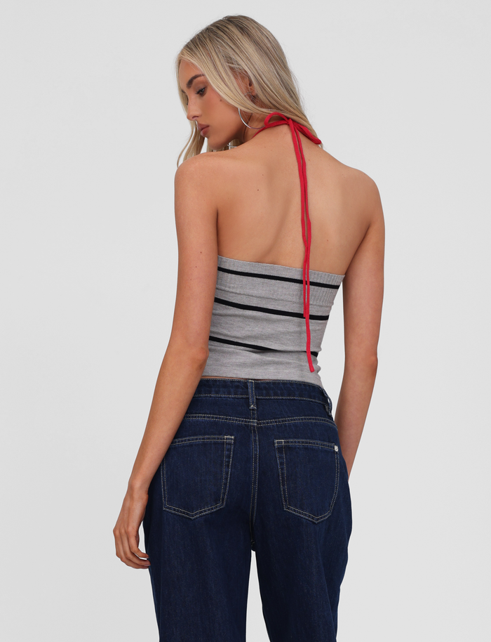 A woman with long blonde hair is seen from the back, wearing the TIGERMIST NOUR TOP - GREY STRIPE with red halter ties and dark blue jeans, standing against a plain light background.