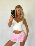 A person with long blonde hair takes a selfie in front of a plain background. They are wearing a white wrap-style crop top from Tiger Mist and the NYDIA SKORT in blush dusty pink by TIGERMIST. They hold a black phone in one hand and pose with the other hand on their hip.