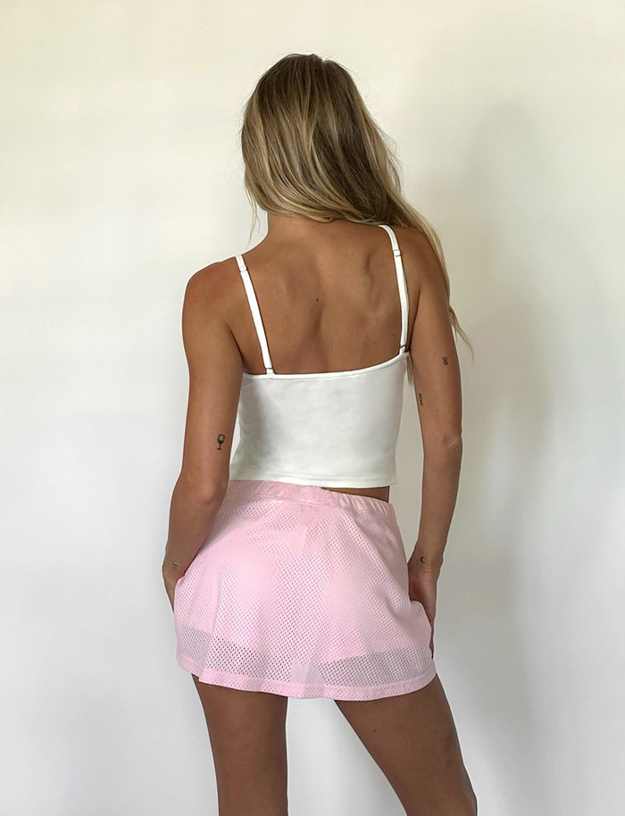 A person with long blonde hair is standing with their back to the camera, wearing a white sleeveless top and a light pink NYDIA SKORT - PINK : BLUSH : DUSTY PINK by TIGERMIST. Their left hand is on their hip, and they are in front of a plain white background.
