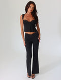 A woman stands against a plain background, wearing TIGERMIST's NYOMI PANT in black with a lace-trimmed top. She has long dark hair and is looking at the camera with a relaxed expression.