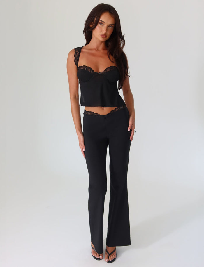 A woman stands against a plain background, wearing TIGERMIST's NYOMI PANT in black with a lace-trimmed top. She has long dark hair and is looking at the camera with a relaxed expression.