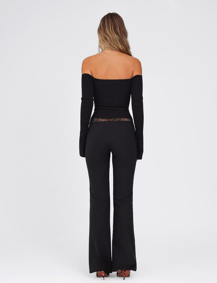 A woman faces away, showcasing the elegant NYOMI PANT - BLACK by TIGERMIST paired with a chic black off-the-shoulder top against a plain white background.