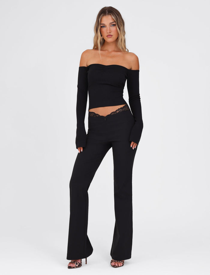 A woman poses against a simple backdrop wearing an elegant black, off-the-shoulder top adorned with intricate lace details and long sleeves. She complements this with the NYOMI PANT in black from TIGERMIST and open-toed heels. Her hands rest by her sides, while her blond hair falls loosely around her shoulders.