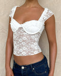 Wearing the TIGERMIST NYRA TOP - WHITE, featuring sheer shoulder straps and floral lace details, paired with blue jeans and set against a light beige textured background.