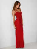 A woman stands against a plain white background wearing the stunning TIGERMIST OCTAVIA MAXI DRESS in cherry red, a long, form-fitting dress adorned with intricate floral embellishments. The elegant design features thin straps, and she poses confidently, her long brown hair cascading over her shoulder.