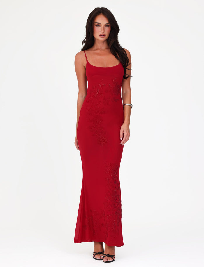 A woman in a sleeveless, cherry red Octavia Maxi Dress by TIGERMIST stands against a white background. She is wearing black high heels and a silver bracelet, with long dark hair cascading over her shoulders. The dress features subtle floral embellishments that add an elegant touch.