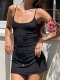 A woman stands in sunlight by a white wall, holding pink flowers, wearing the TIGERMIST OCTAVIA MINI DRESS - BLACK with adjustable spaghetti straps and floral embellishments. Her face is not visible.