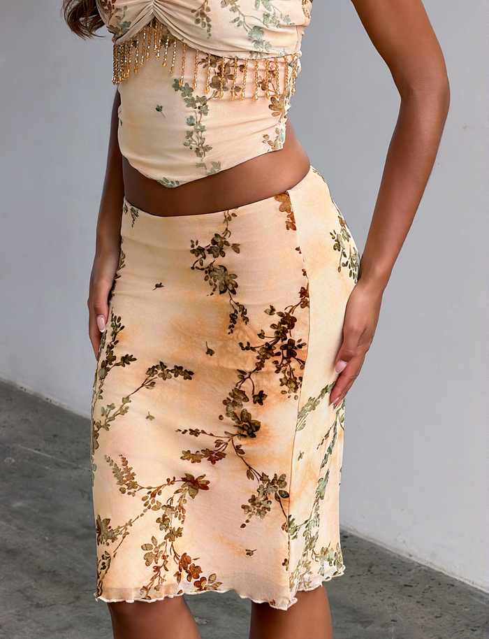 A woman stands by a light-colored wall wearing the TIGERMIST OONA BURN OUT FRINGE SKIRT in tan, paired with a matching sleeveless crop top featuring bead details.
