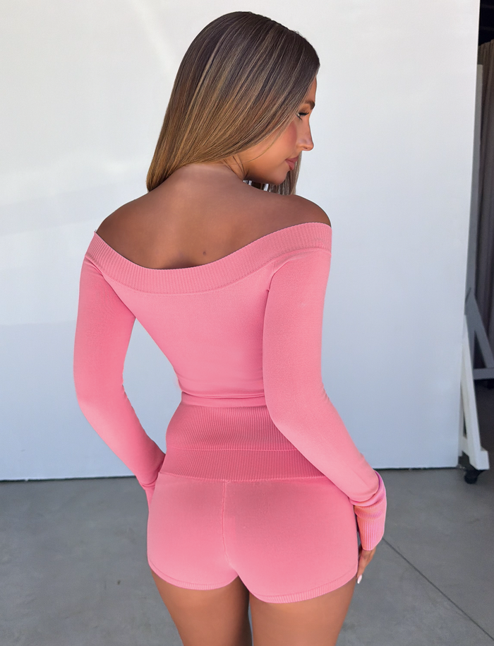 A woman with long, straight hair stands facing away, wearing a fitted off-the-shoulder pink top paired with the TIGERMIST PARKER KNIT MICRO SHORT in Coral, which features a fold-over ribbed waistband, set against a plain light-colored background.