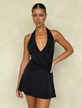 A woman confidently poses against a plain light-colored background, wearing a sleeveless PARRIS MINI DRESS - BLACK from TIGERMIST, featuring a deep V-neckline and a knot detail, with her hand on her hip as she gazes at the camera.