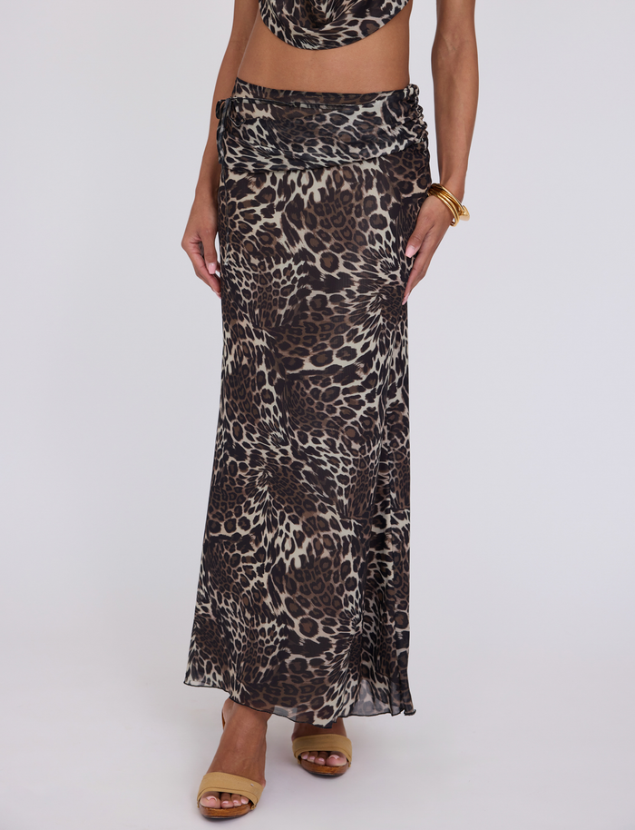 A person wears the TIGERMIST PEARL MAXI SKIRT - MULTI : LEOPARD, featuring a long, fitted silhouette with a ruched waist. Styled with brown sandals and gold bracelets, only the lower body is visible against a plain white background.