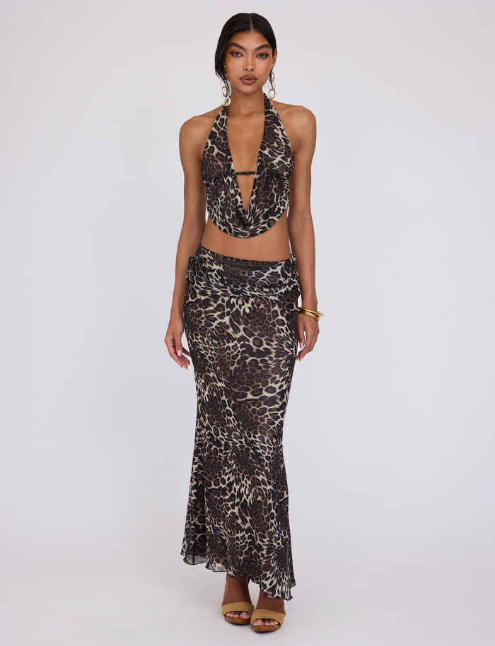 A woman poses against a plain background in the TIGERMIST PEARL TOP - MULTI: LEOPARD, a sleeveless halter with an open back and plunging neckline, styled with a long skirt, gold earrings, bracelets, and brown heels.