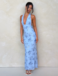 A woman poses against a beige wall in the TIGERMIST PHAEDRA MAXI DRESS - BLUE FLORAL, a floor-length halter dress with a deep V-neck and low open back. She pairs it with white heeled sandals and wears her dark hair up.