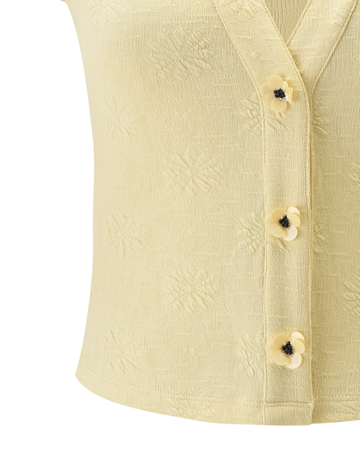 The TIGERMIST QUINLAN TOP in Lemon is a light yellow, sleeveless, stretchy top with a V-neck and four floral-shaped buttons with dark centers, featuring subtle woven and floral details for added style.