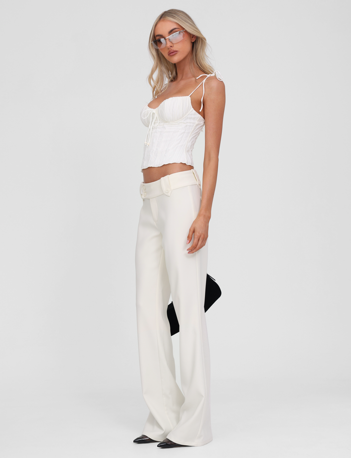 A woman stands against a plain white background wearing the TIGERMIST RANIA TOP in off white, styled with high-waisted wide-leg white pants, black pointy shoes, glasses, and holding a black clutch behind her.