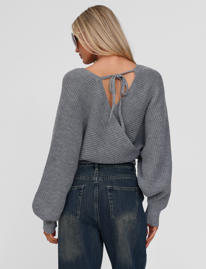 A woman with long blonde hair wears sunglasses, high-waisted blue jeans, and the TIGERMIST RAVEN JUMPER in grey featuring a crisscross back and tie detail. She faces away from the camera against a plain background.