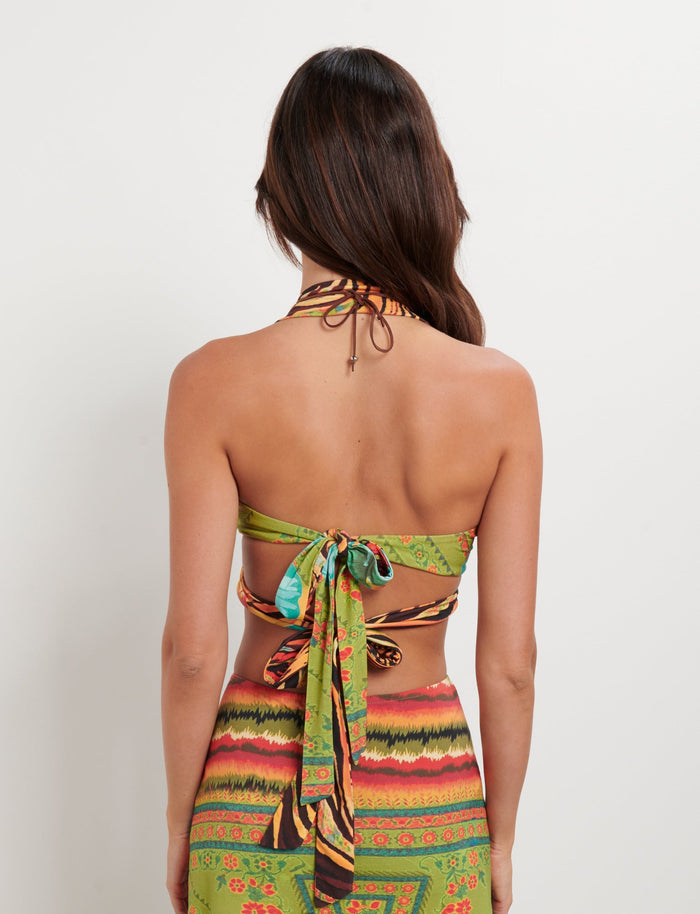 A woman with long dark hair stands with her back to the camera, showcasing a vibrant TIGERMIST RAYLEE TOP - MULTI : FLORAL : TROPICAL FLORAL halter-neck top that ties in a bow at the back, paired with a matching skirt featuring bright green, red, and yellow patterns.