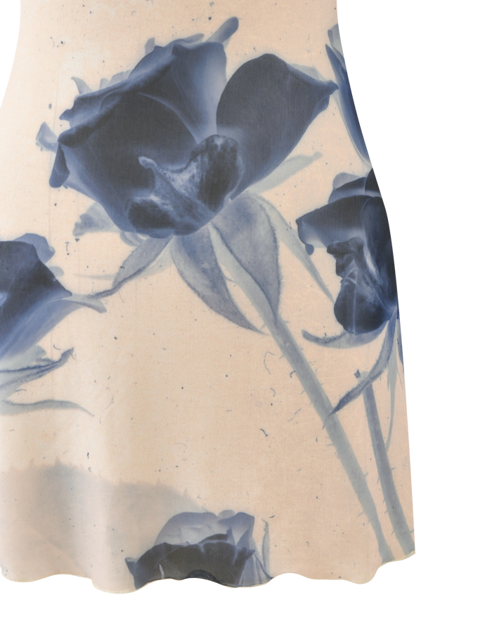 The ROSY MINI DRESS - INDIGO FLORAL by TIGERMIST is a light beige mini dress with adjustable cami straps, showcasing large artistic blue rose prints with stems and leaves that pop against the plain background.
