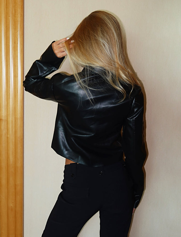 A person with long blonde hair stands with their back to the camera, wearing the stylish TIGERMIST RYDER JACKET - BLACK and black pants. Their left hand is touching their hair as they stand against a light-colored wall with wooden trim.