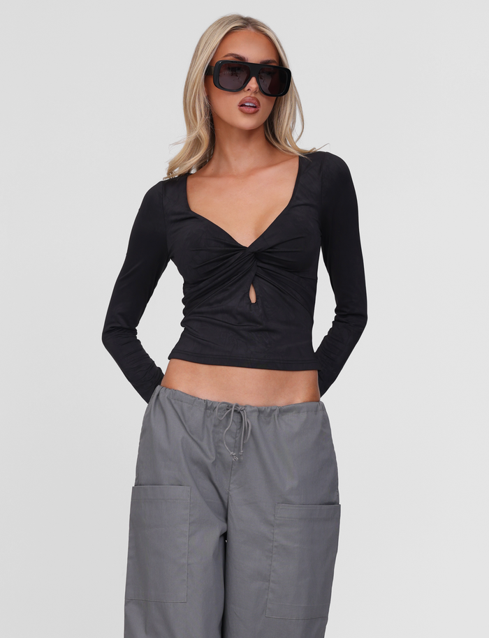 A woman wears the TIGERMIST SAFFIRA LONG SLEEVE TOP in black, featuring a twist front and keyhole V neckline, paired with loose gray pants and oversized sunglasses as she poses against a plain white background.