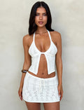 A woman with long dark hair poses in front of a neutral background. She is wearing a white crocheted halter top with a front opening and the SANDOR SKIRT - WHITE from TIGERMIST, featuring a drawstring waist. She has a bracelet on her right wrist.